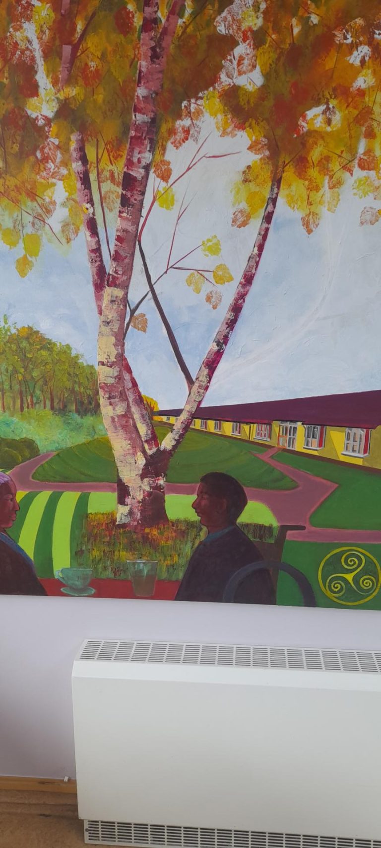 Birr Art Group presented a mural to commemorate 20 years of the Sandymount Nursing Unit.  Kathleen Gormley & Jean Forristal were key to the project.  The triskel in the bottom right corner with its three spirals represents the cycle of birth, life and death.  It is also a symbol of strength and progress.  The Nursing home and grounds are a central feature to the Mural. While two residents sit around the drumlin of tree shrubs.  The mural is displayed in the day care center activity room.